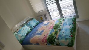 A bed with a colorful landscape design, a window with blinds, and a visible air conditioning unit. - 2 BR Condo by JAD at Azure Urban Resort Residences (Parañaque)