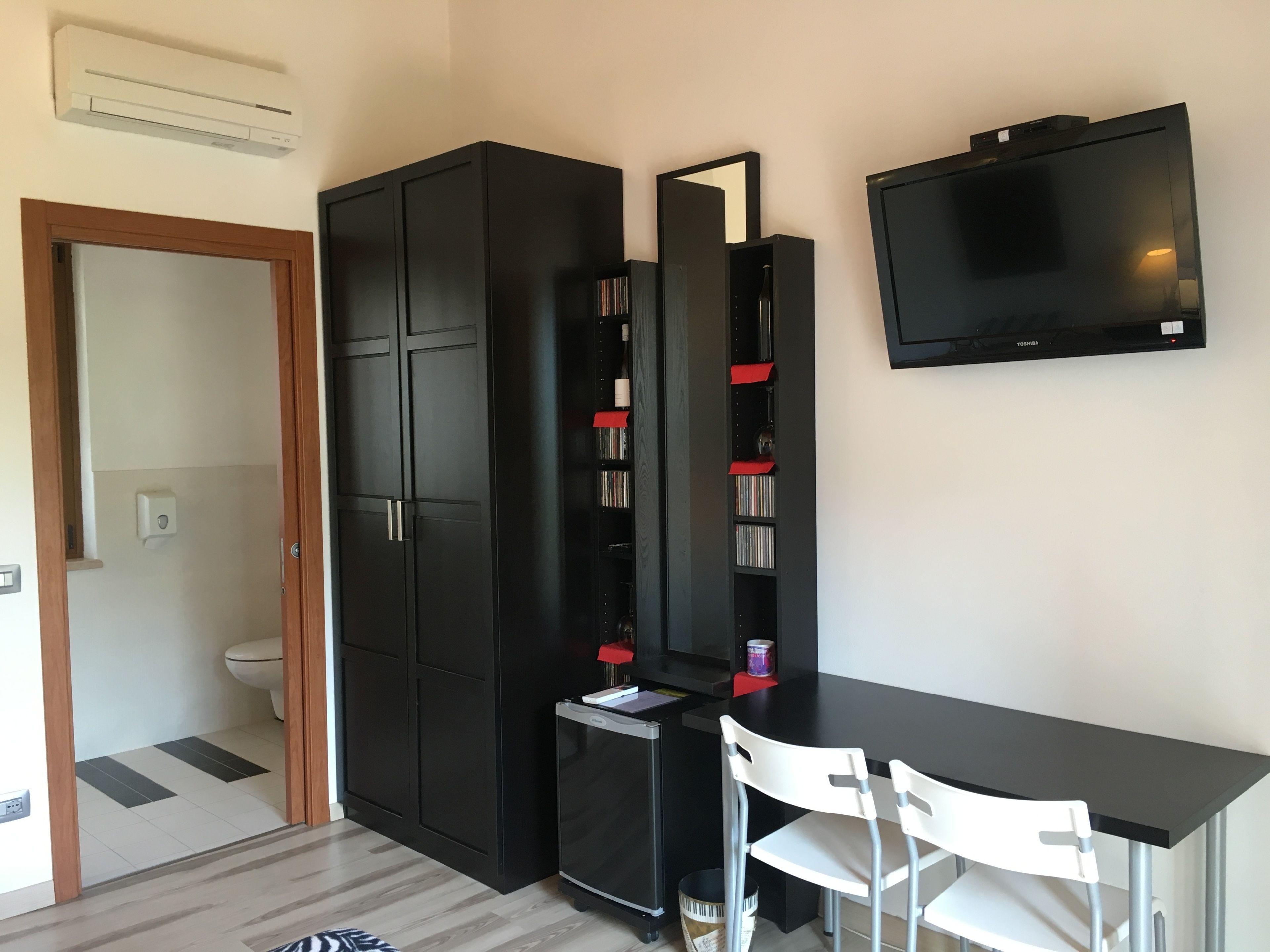Une pièce de taille modeste, avec une armoire noire, une petite table accompagnée d’une chaise blanche, un téléviseur fixé au mur et un miroir.