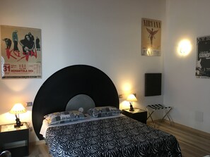 Quarto com uma cama grande, duas mesas de cabeceira com abajures, uma cadeira e cartazes na parede.