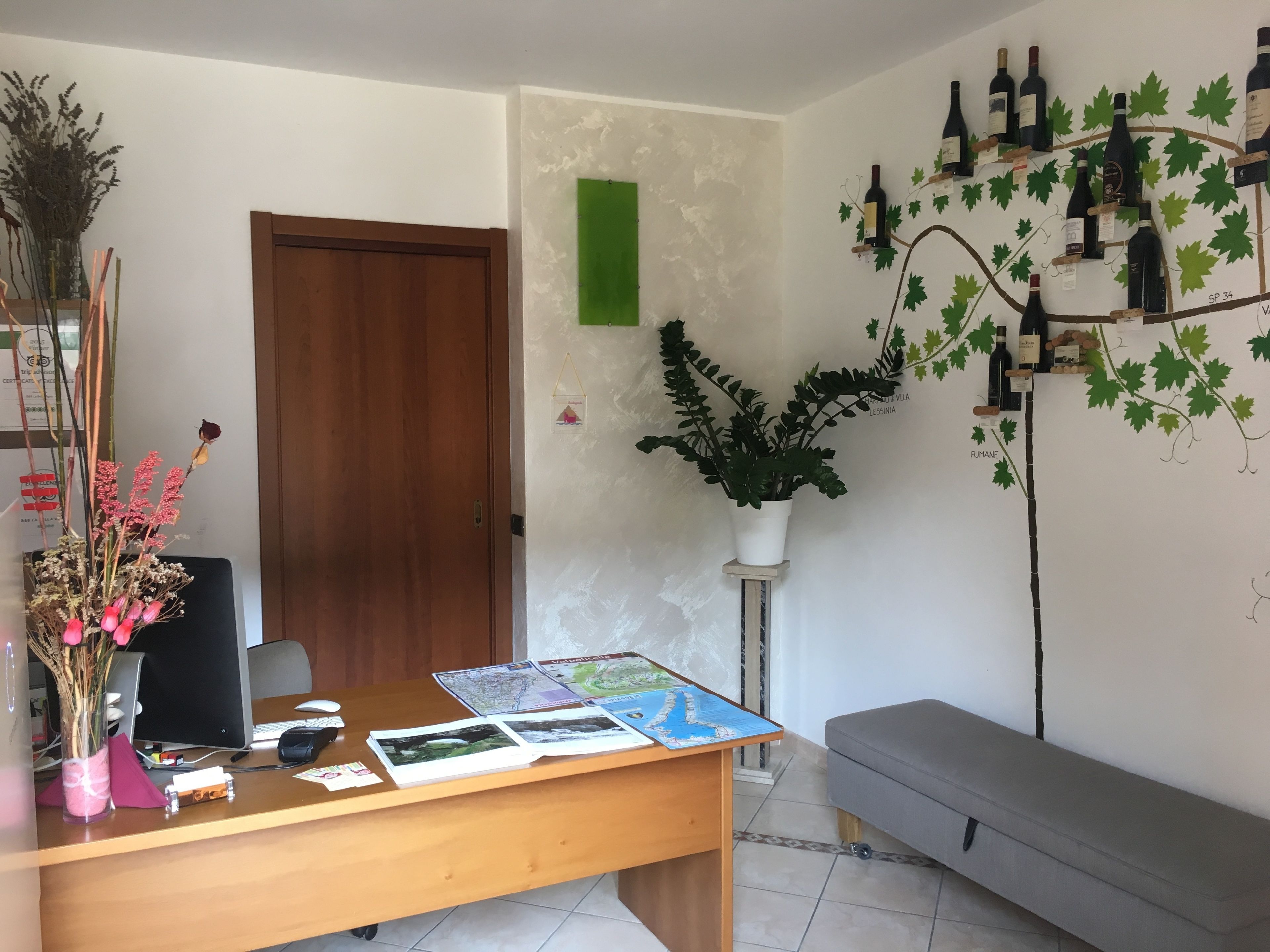 A reception desk with a computer, papers, and a map. There is a potted plant, a wooden door, and a wall with wine bottles and leaves.