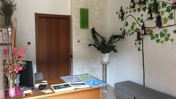 A reception desk with a computer, papers, and a map. There is a potted plant, a wooden door, and a wall with wine bottles and leaves.