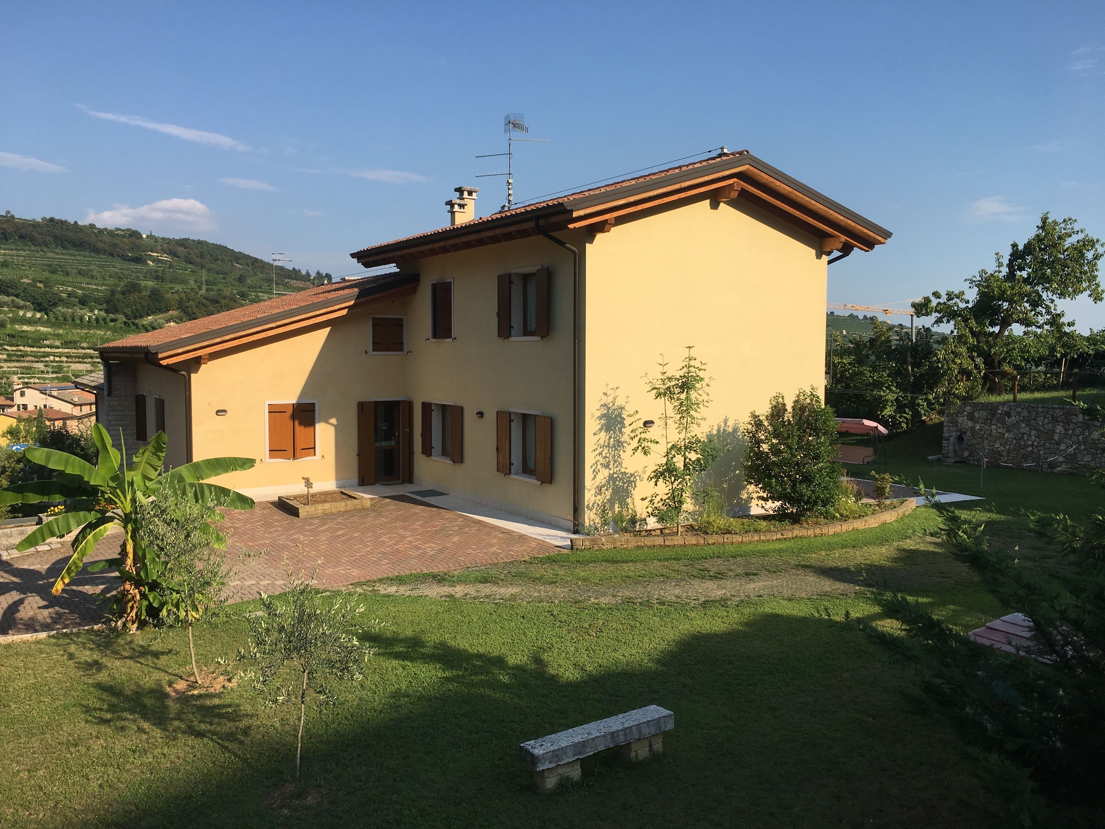 A two-story house with a tiled roof, a stone wall, and a small garden area with a bench.