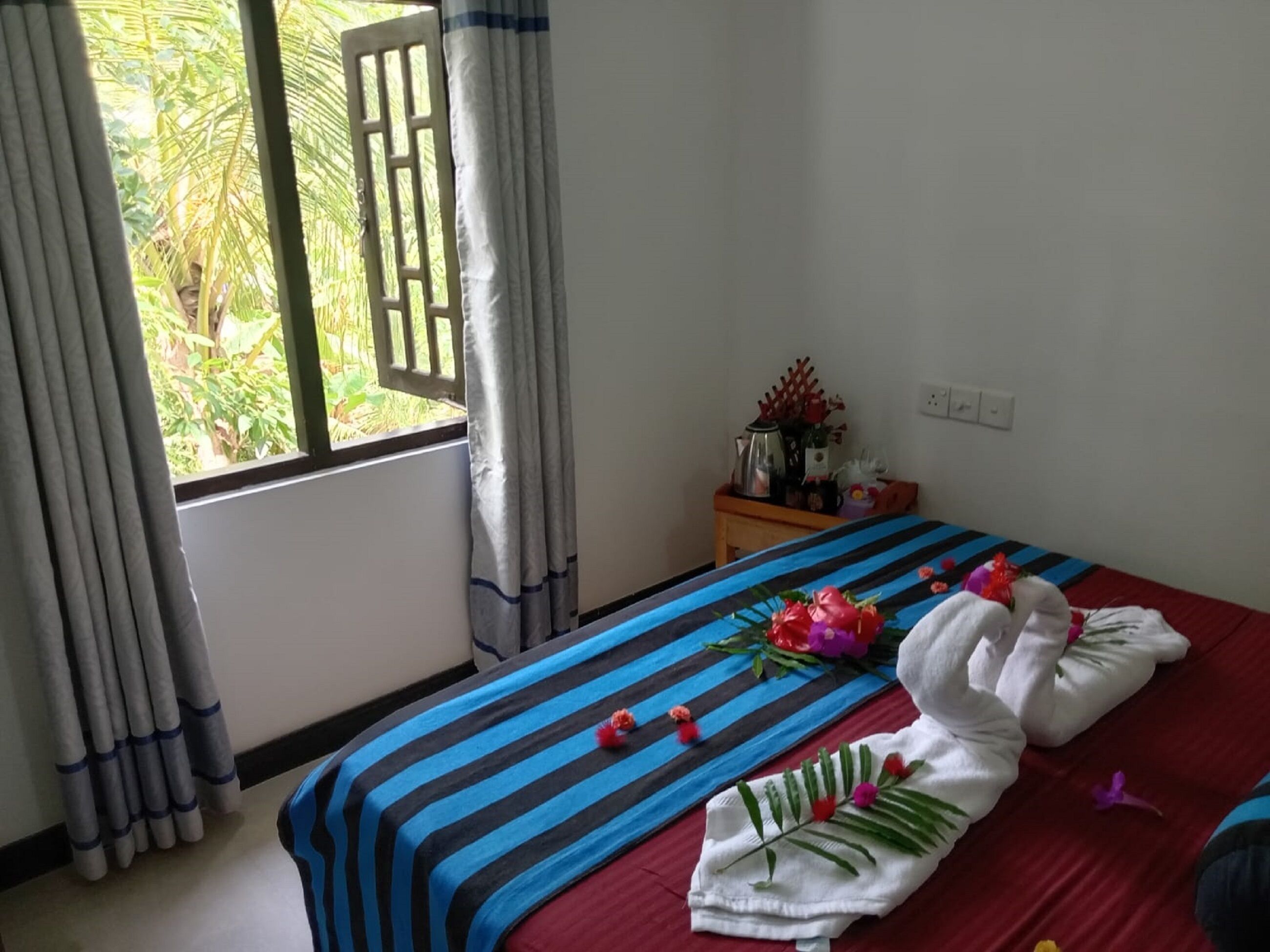 A hotel room with a bed, a window with curtains, a nightstand with a lamp, and a view of greenery outside.