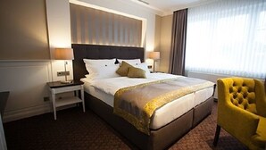 Een hotelkamer met een groot bed, twee nachtkastjes, een gele fauteuil en een raam met gordijnen.
