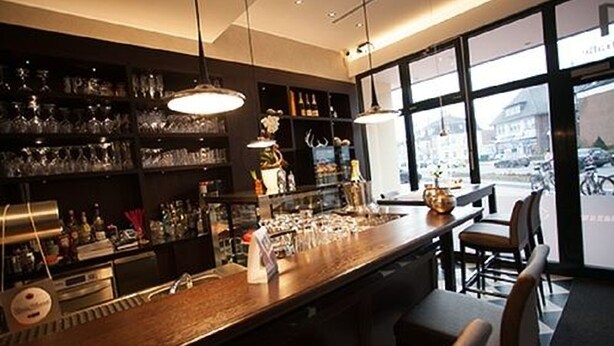 Een bar met glazen en flessen op planken, een bargedeelte en uitzicht op de straat door grote ramen.