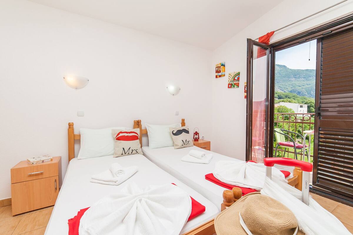 Un lit double avec du linge de lit blanc et des touches de rouge, une table de chevet en bois avec un livre, un chapeau rouge et un balcon donnant sur un paysage verdoyant.