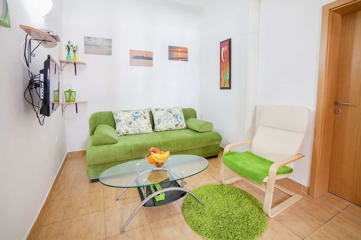 Un petit salon avec un canapé vert, une table basse en verre, un fauteuil blanc et un tapis vert.