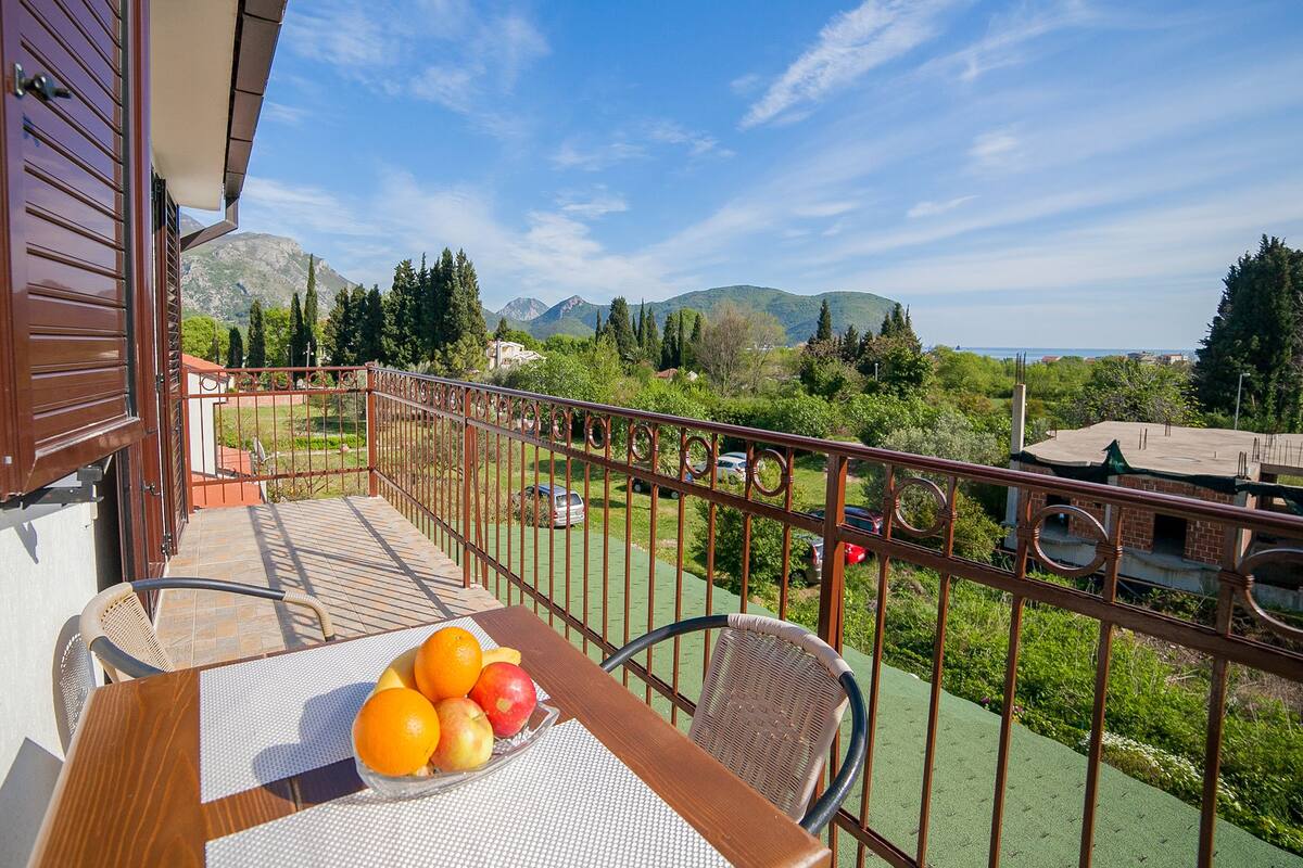 Un balcon avec une table dressée, ornée de fruits, donnant sur un paysage verdoyant avec des montagnes et des voitures garées.