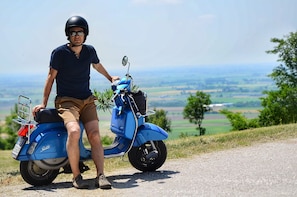 Una persona sentada en un patinete azul por un camino rural, con una vista panorámica de campos y árboles al fondo.