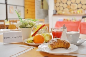 Una mesa con desayuno que incluye un croissant, frutas, jugo y café, con un cartel que dice "CASTEL SAN ZENO".