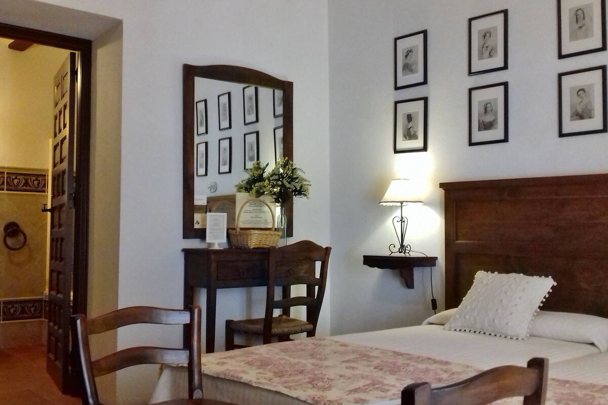 Una habitación de hotel con una cama, un escritorio con lámpara, una silla, un espejo y cuadros en la pared.