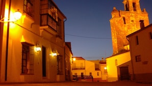 Un edificio histórico con un campanario, iluminado por una luz cálida, y una calle con un banco al frente.