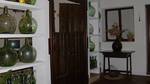 Una habitación con estantes que exhiben varias botellas de vidrio verde, una puerta de madera, una ventana con vista a una planta y una pequeña mesa con un florero.