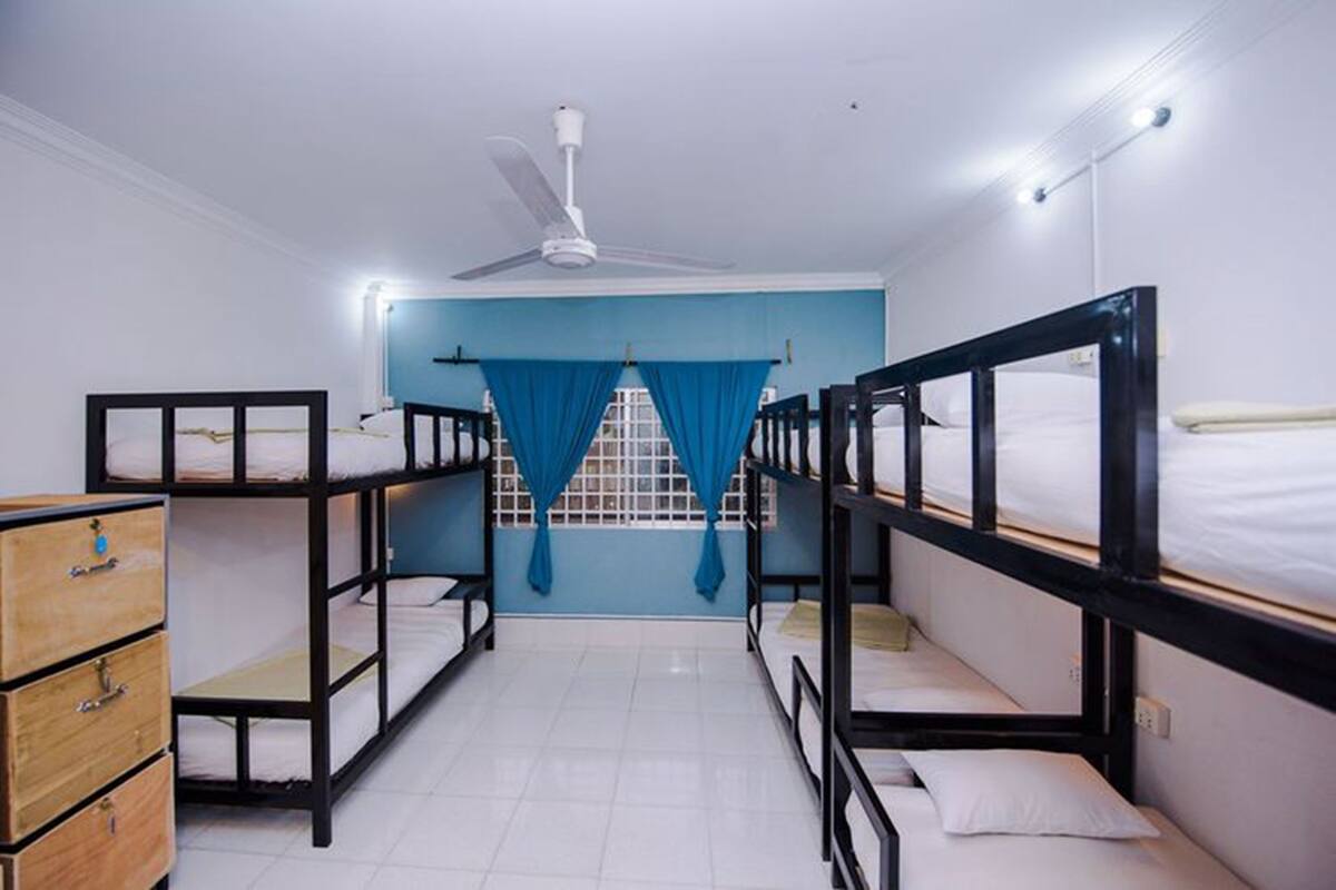 Um dormitório com beliches, um ventilador de teto, uma janela com cortinas azuis e uma cômoda de madeira.