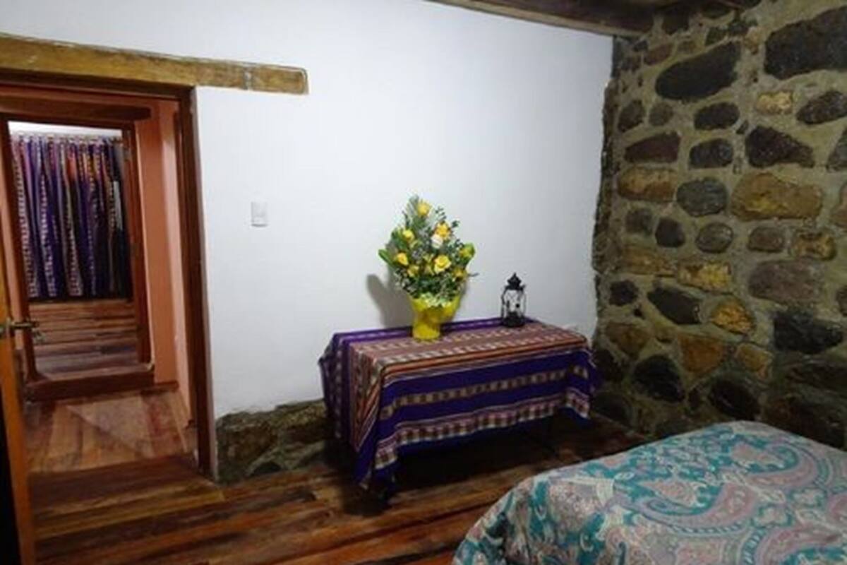 A room with a wooden door, a small table with a patterned tablecloth, a vase with flowers, and a bed with a patterned bedspread.