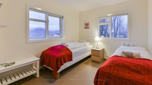 Ein Schlafzimmer mit zwei Betten, einem Nachttisch, einem Regal und zwei Fenstern mit Blick auf eine verschneite Landschaft.