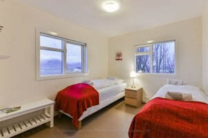 A bedroom with two beds, a nightstand, a shelf, and two windows with views of a snowy landscape.