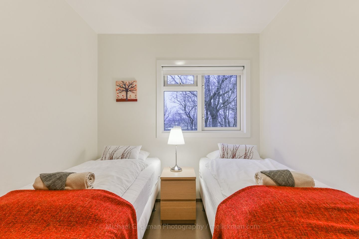 Une chambre avec deux lits, une table de chevet, une lampe et une fenêtre donnant sur des arbres sans feuilles.