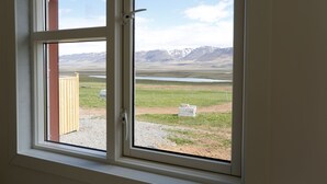 Ein Fenster mit Blick auf ein grasbewachsenes Feld, eine Holzhütte und ferne Berge.