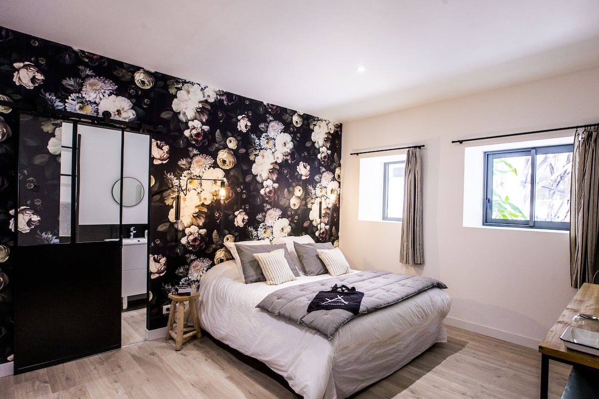 A bedroom with a floral wallpaper, a bed with multiple pillows, a bedside table with a lamp, and a window with curtains.
