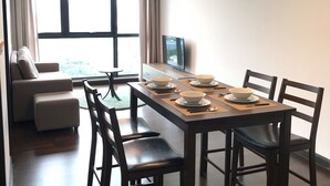 Un comedor con una mesa larga dispuesta para cuatro personas, rodeada de sillas, con un sofá y un televisor al fondo.