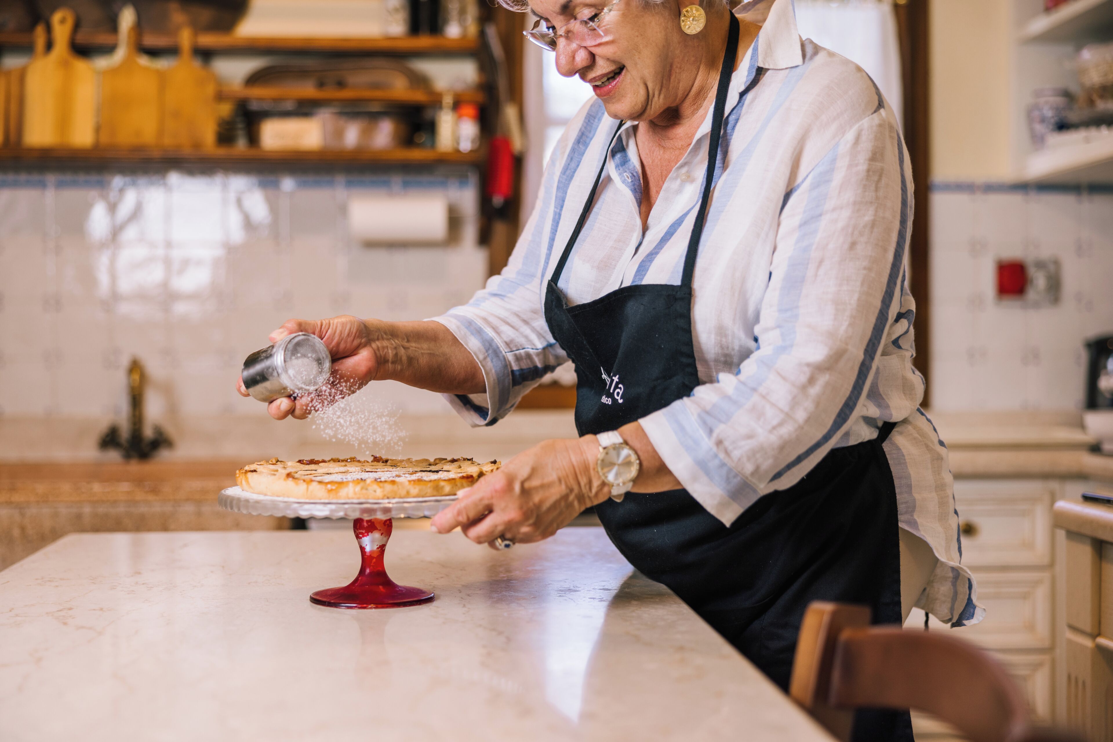 Eine Person in einer Küche, mit Schürze bekleidet, streut eine pulverförmige Substanz auf einen Kuchen auf einem roten Sockel.