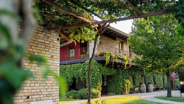 Un edificio de ladrillo con puerta roja, un balcón con plantas y un jardín con mesas y sillas.