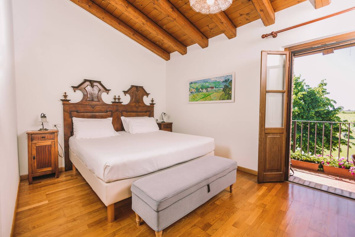 Un dormitorio con techo de madera, cama con ropa de cama blanca, mesita de noche, banqueta y balcón con plantas en macetas.
