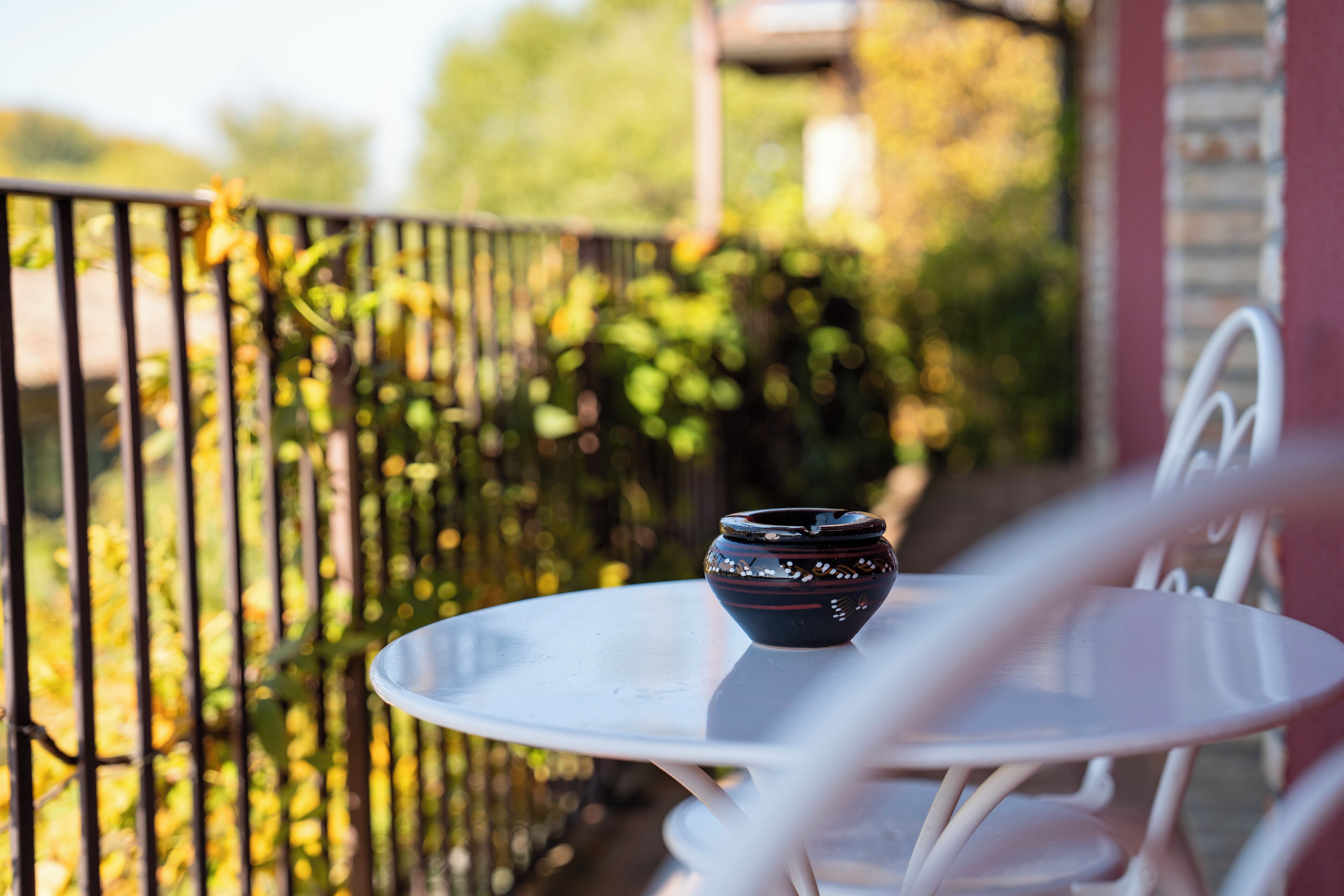 A white metal table with a ceramic bowl on it, set on a balcony with a metal railing and greenery in the background.
