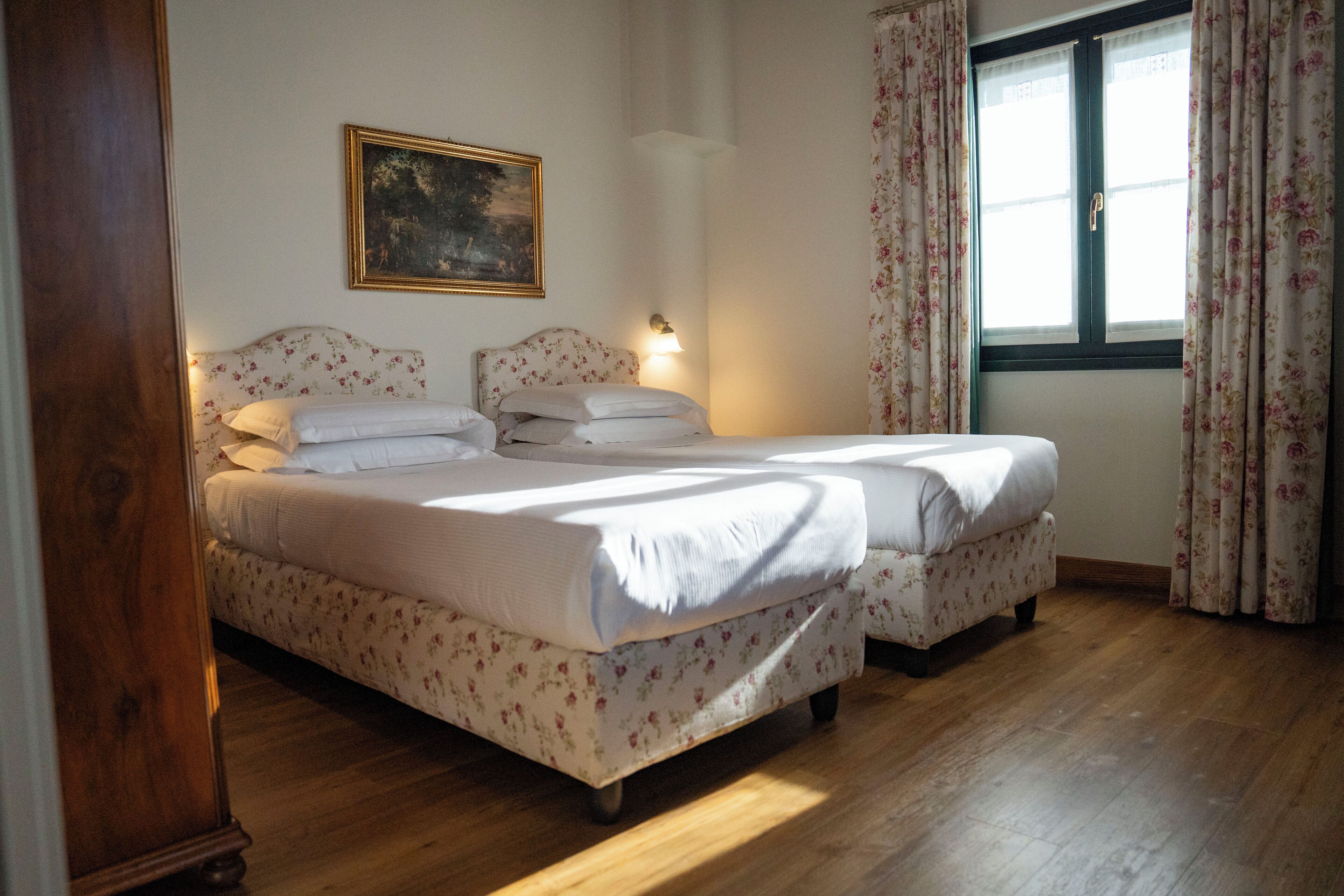 A double bed with floral bedding, a nightstand, a wall-mounted lamp, and a window with floral curtains.