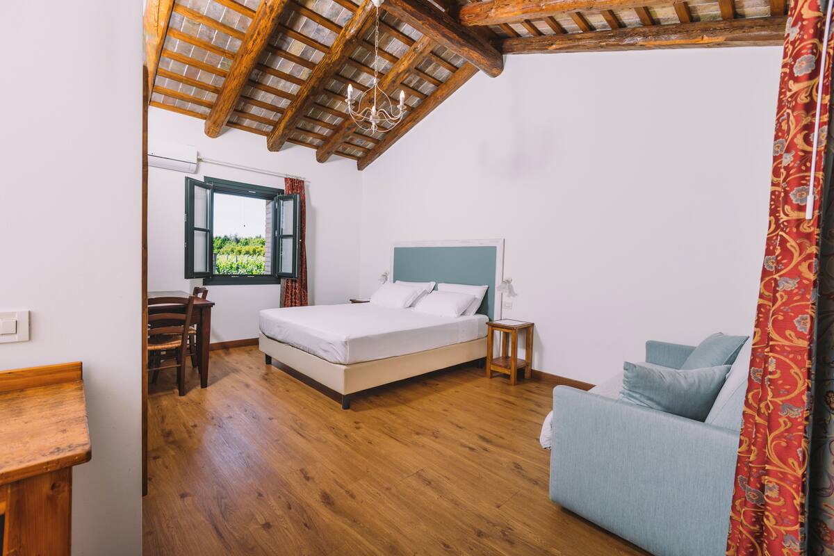 Un dormitorio con techo de madera, cama con ropa de cama blanca, mesita de noche pequeña, ventana con vistas a zonas verdes y un sofá azul.