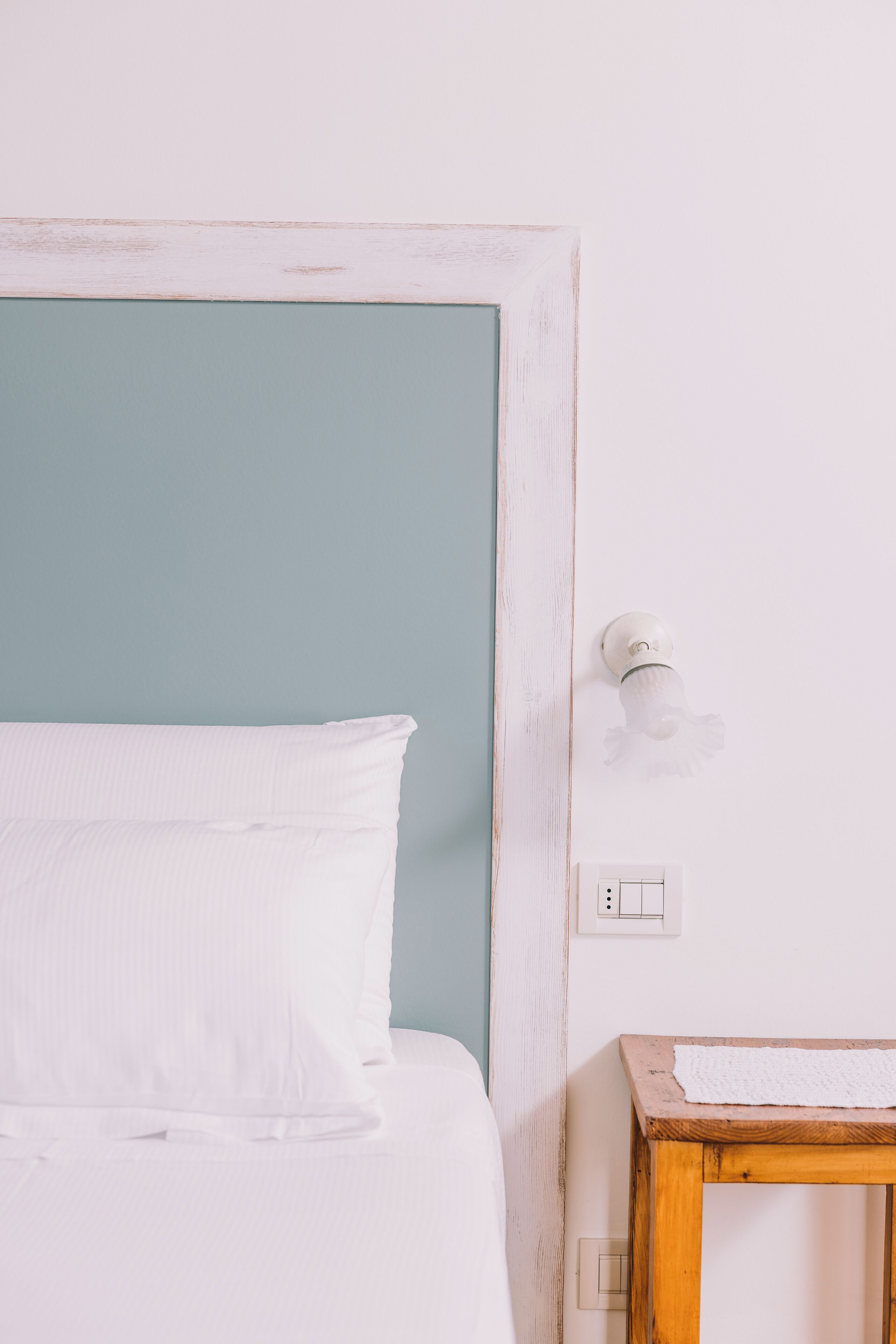 A bed with white bedding, a wooden nightstand with a white cloth, and a wall socket.