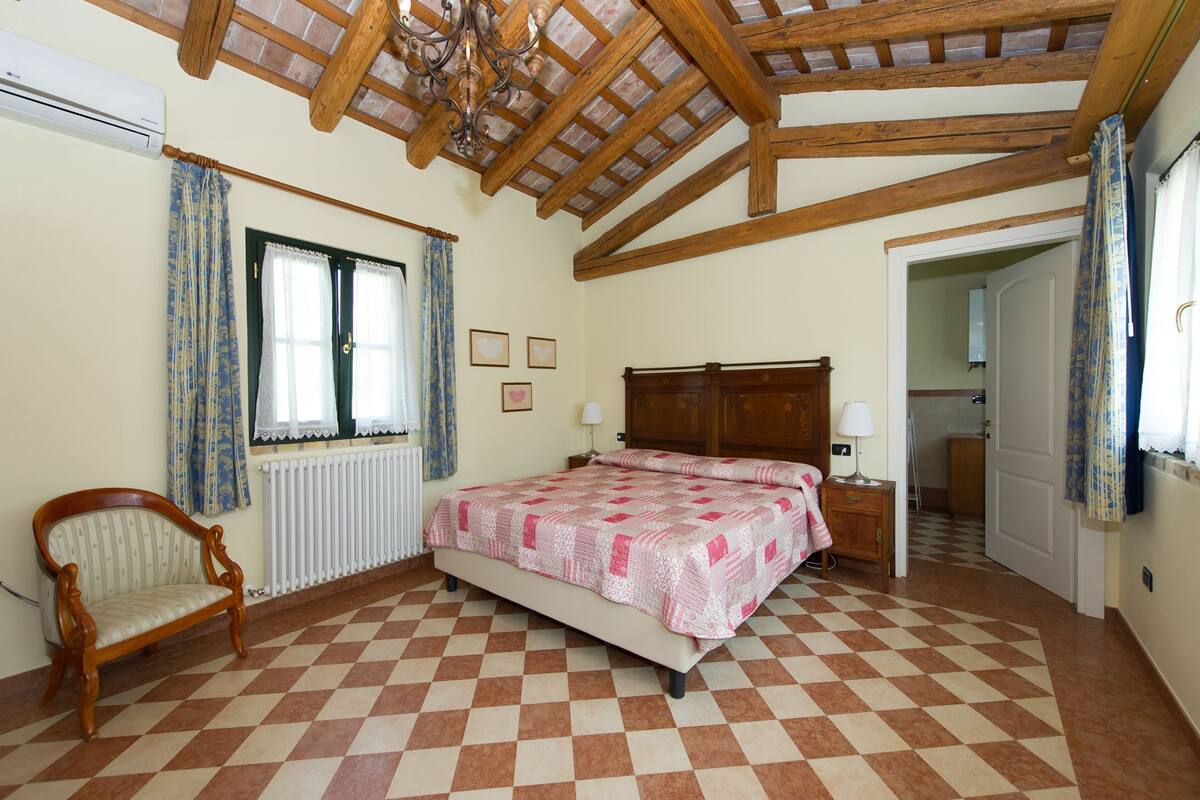 Un dormitorio con suelo a cuadros, una cama de madera, una silla, un radiador y un techo con vigas a la vista.
