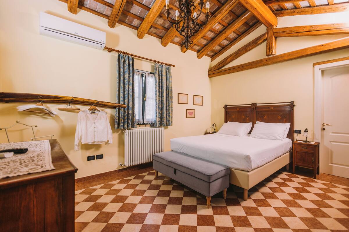 Un dormitorio con suelo a cuadros, una cama, una mesita de noche, un aparador, un perchero y una ventana con cortinas.