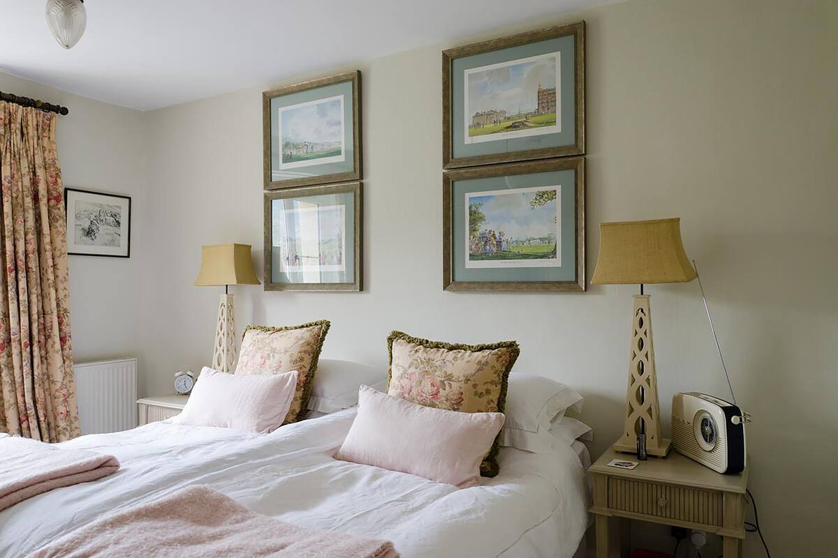 A neatly made bed with floral pillows, a bedside table with a lamp and a vintage radio, and three framed pictures on the wall.