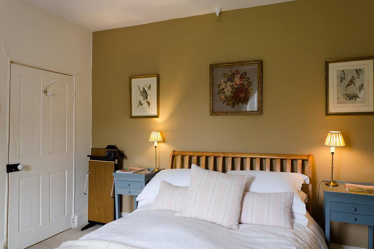 A bedroom with a wooden bed, two bedside tables with lamps, a nightstand, a door, and three framed pictures on the wall.