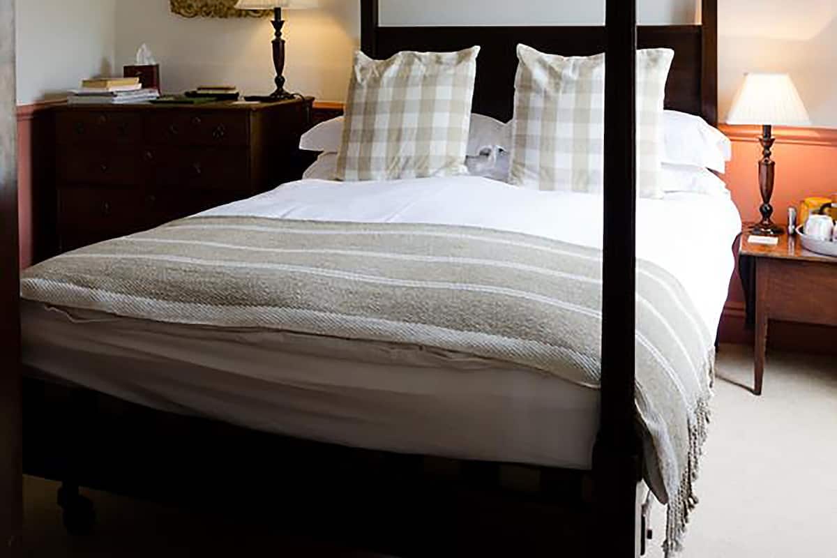 A four-poster bed with a white bedspread and checkered pillows, a wooden nightstand with a lamp, a mirror with an ornate frame, and a framed picture on the wall.