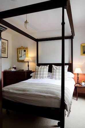 A four-poster bed with a white bedspread and checkered pillows, a wooden nightstand with a lamp, a mirror with an ornate frame, and a framed picture on the wall. - Heron House (Tiverton)