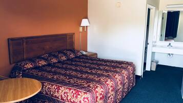 Quarto de hotel com uma cama grande, mesa de cabeceira, abajur, uma pequena mesa e banheiro com pia e espelho.