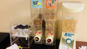 A dispenser with Raisin Bran and Froot Loops cereals, a container of General Mills cereal, and a bowl of bananas.