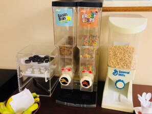 A dispenser with Raisin Bran and Froot Loops cereals, a container of General Mills cereal, and a bowl of bananas.