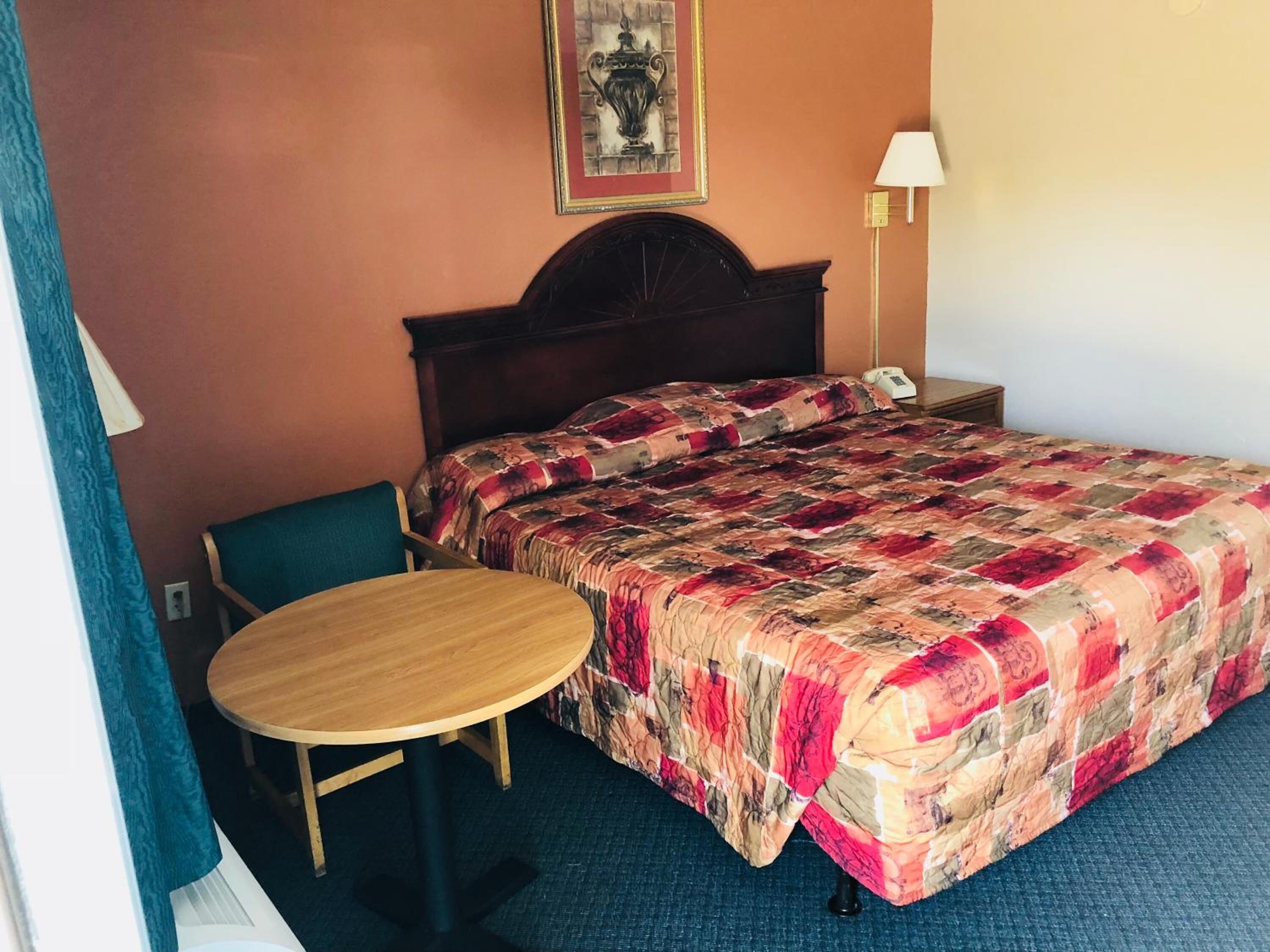 A hotel room with a bed, a round wooden table, a chair, a nightstand with a lamp, and a painting on the wall.