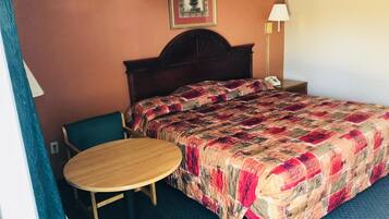 A hotel room with a bed, a round wooden table, a chair, a nightstand with a lamp, and a painting on the wall.