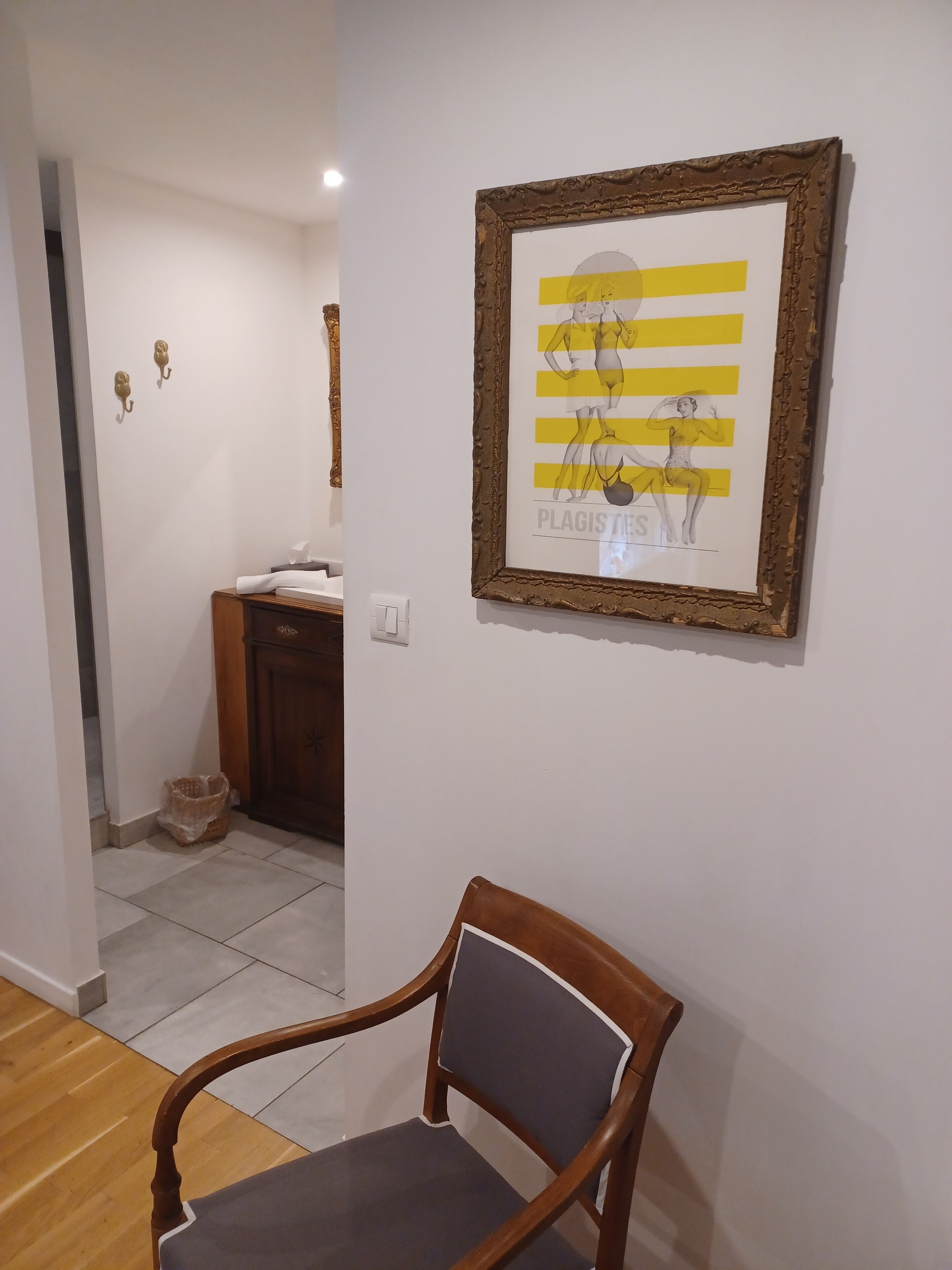 Een kamer met een houten stoel, een kunstwerk in een lijst aan de muur, een houten kast en een kleine prullenbak.