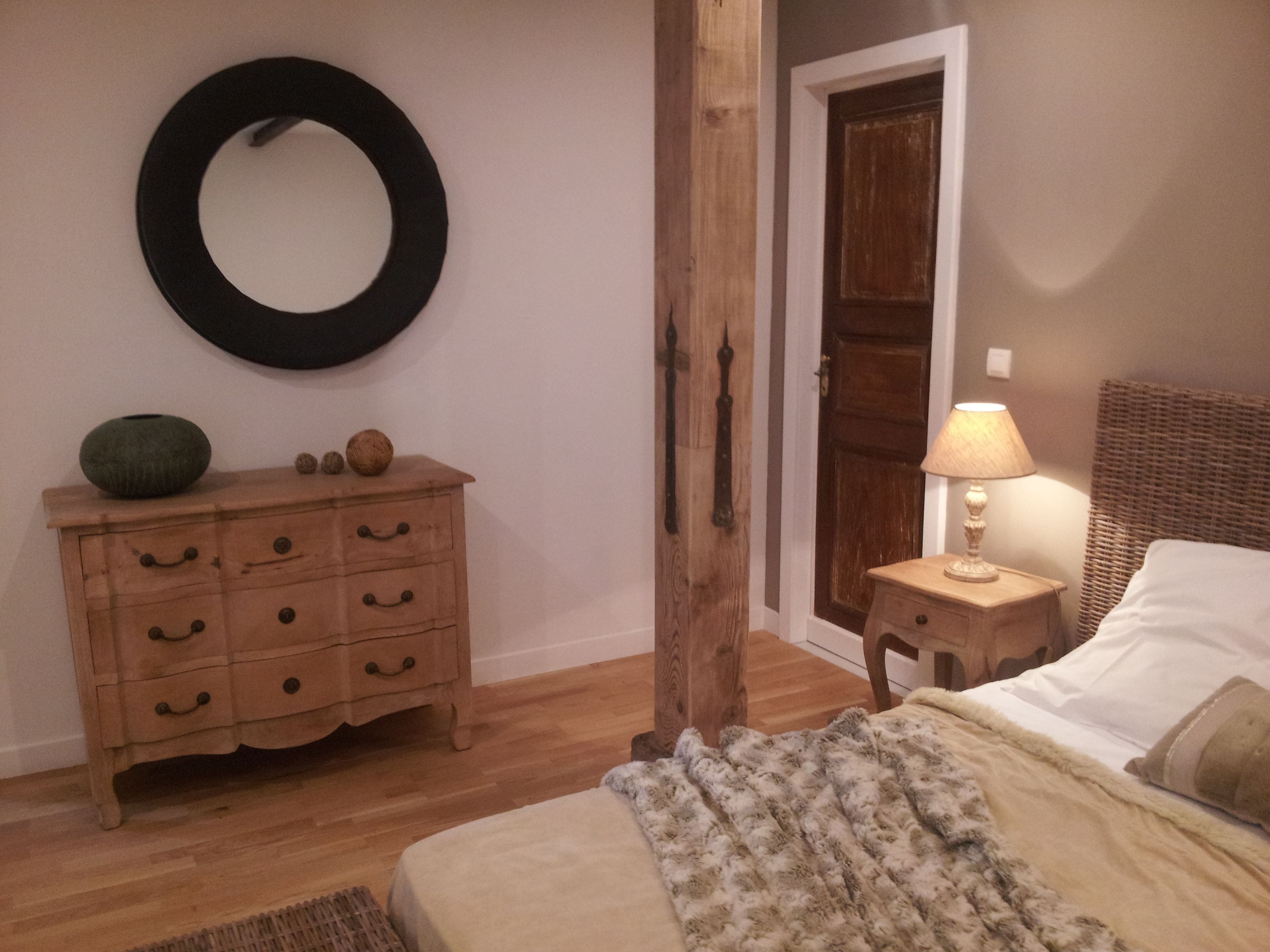 Ein Schlafzimmer mit einem Holzkonsolen, einem runden Spiegel, einer Tür, einem Nachttisch mit Lampe und einem Bett mit Decke.