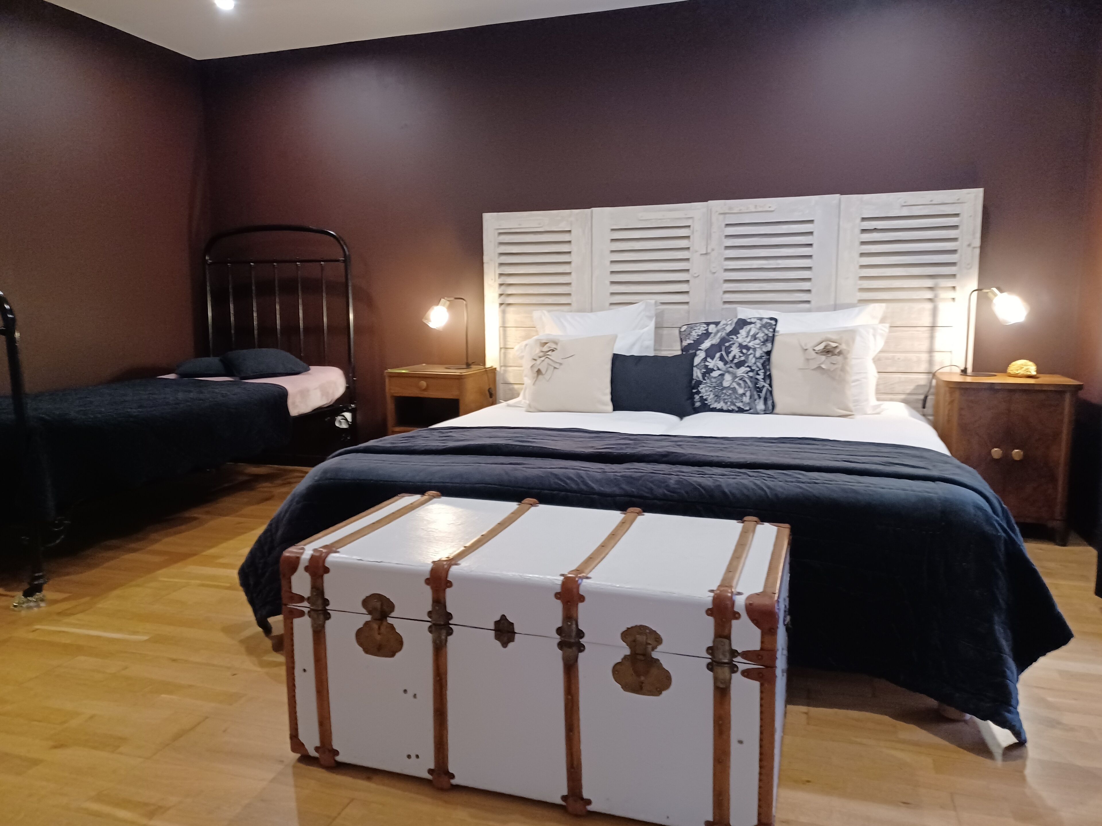 Un dormitorio con una cama grande, una cama más pequeña, una mesita de noche, una lámpara y una maleta.