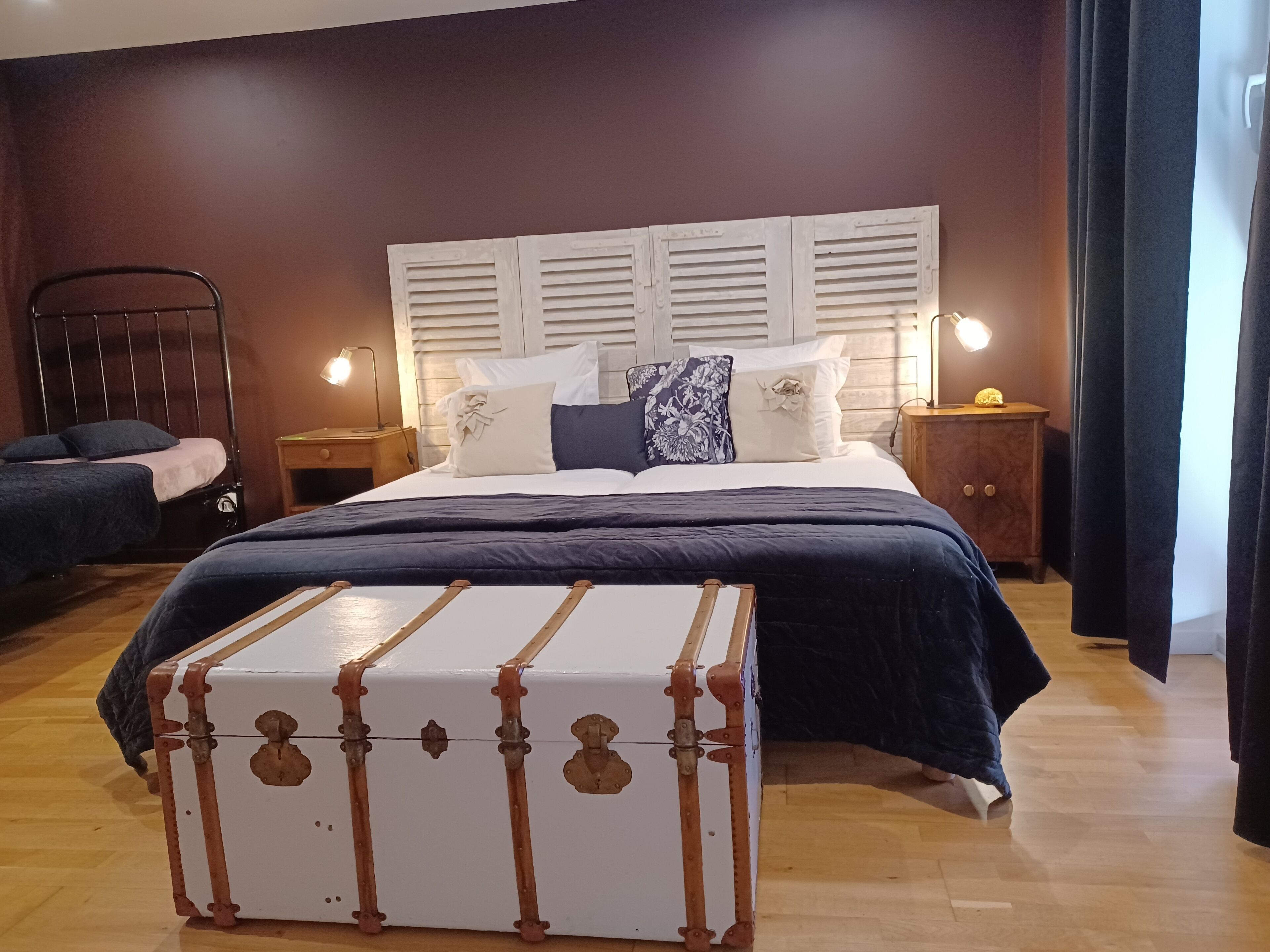 A bedroom with a large bed, two bedside tables, a nightstand, a wooden floor, and a vintage suitcase in the center.