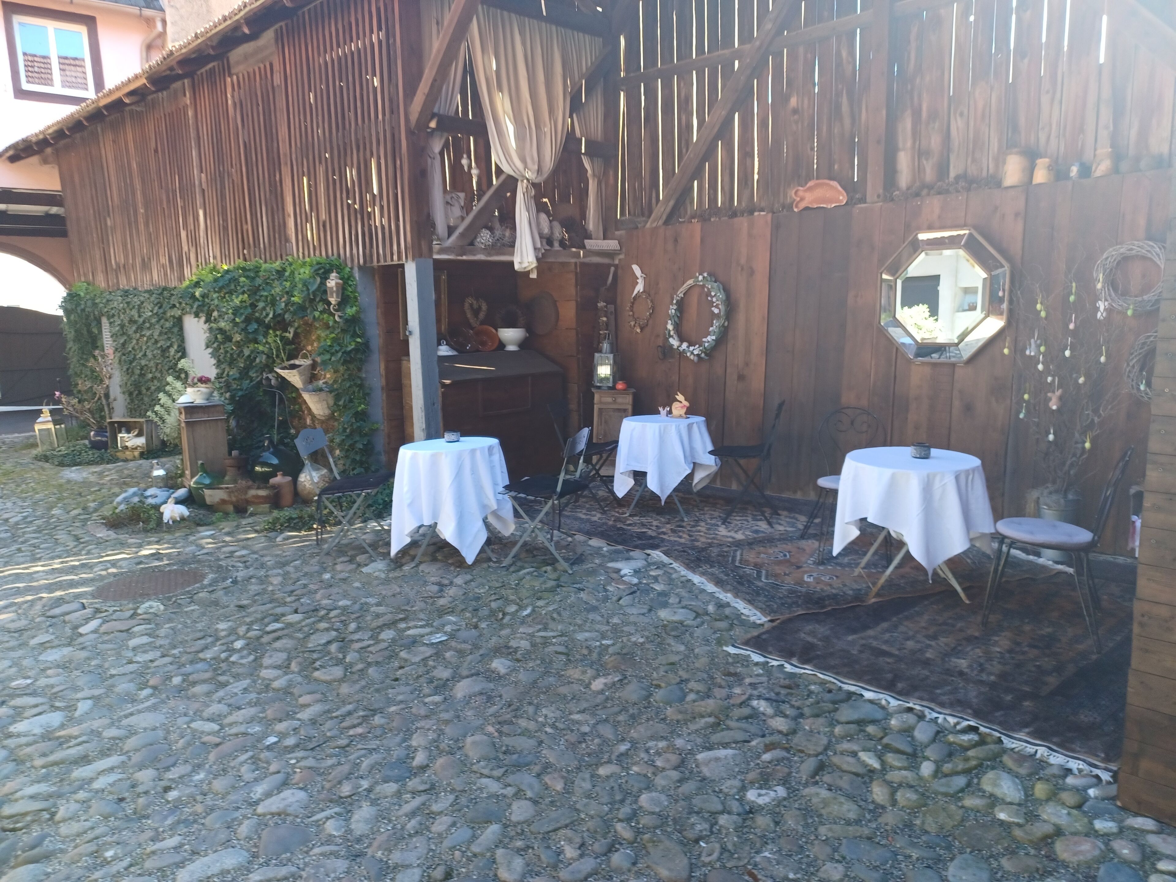 An outdoor seating area with tables and chairs, a wooden building with a sloped roof, and decorative elements like wreaths and lanterns.