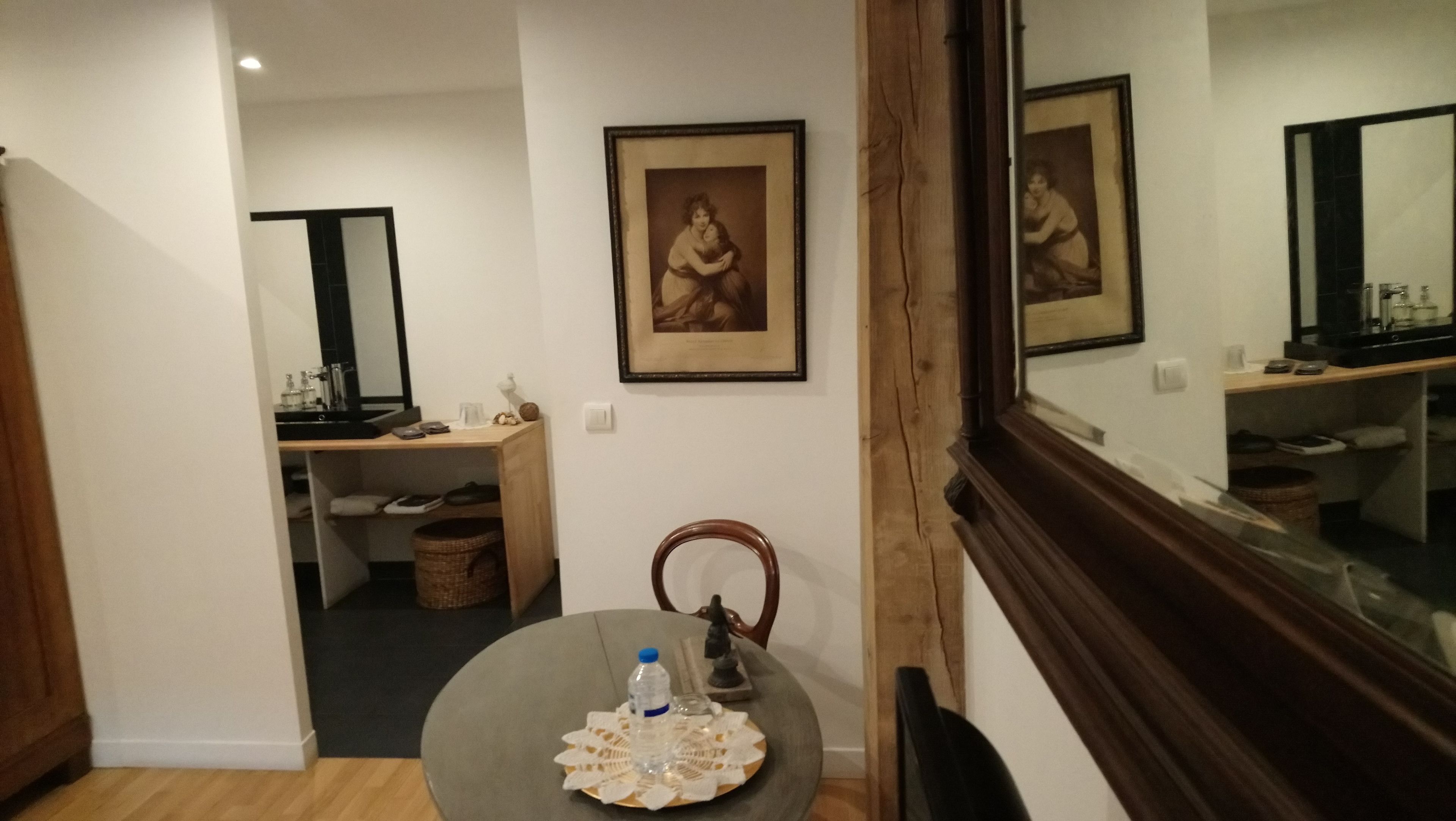 Ett hotellrum med trägolv, ett runt bord med en vattenflaska och en spetsduk, en trästol, en liten statyett, en inramad tavla på väggen och en spegel som reflekterar en del av rummet.