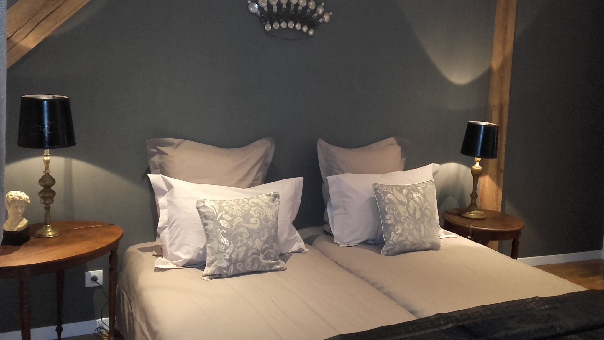 Cama doble con dos almohadas blancas y un cabecero decorativo. Hay dos mesitas de noche, una con lámpara y una escultura de busto.